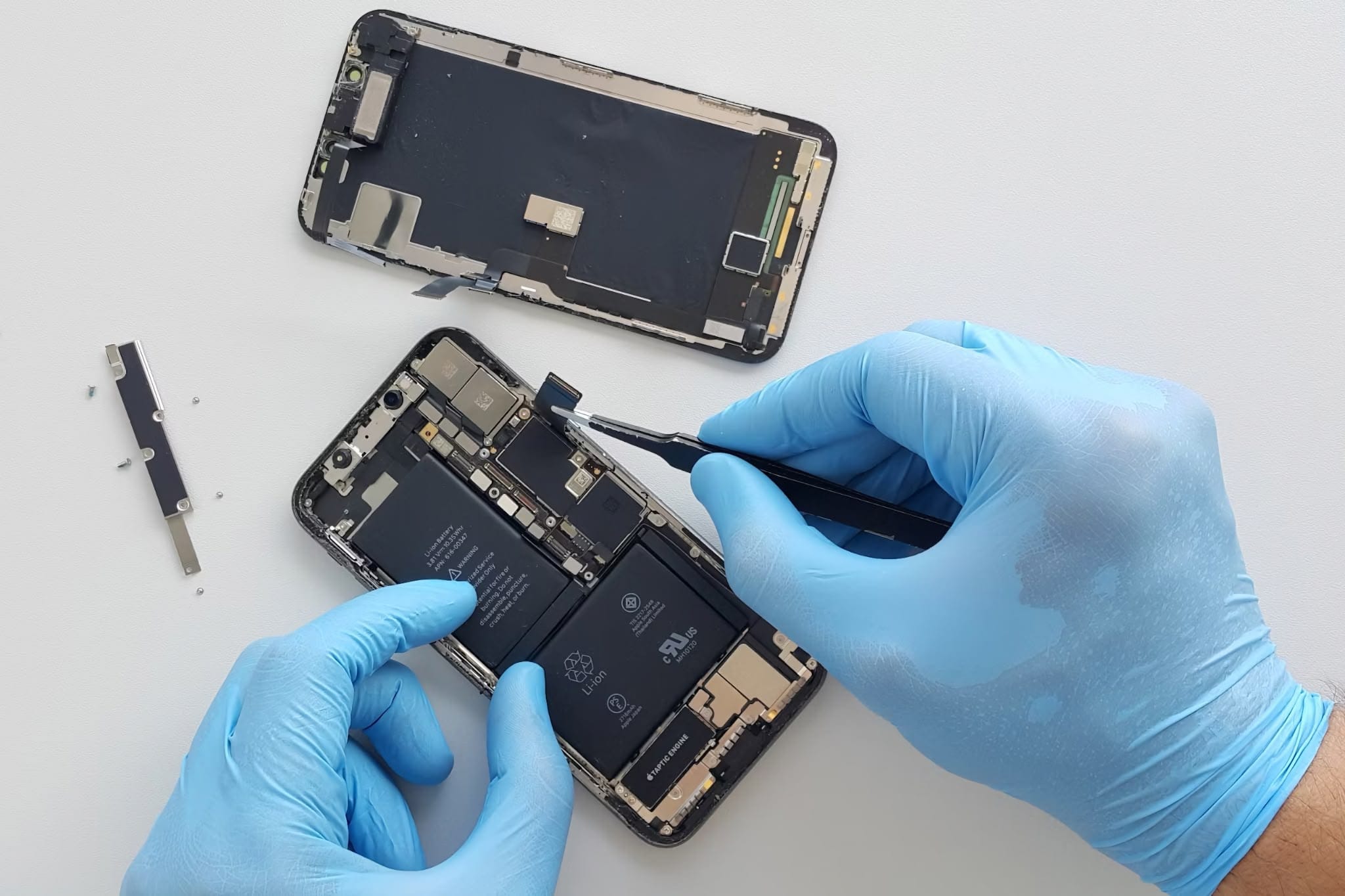 Gloved hands repair an opened smartphone on a white surface: the display lies face‑down above while tweezers lift a ribbon connector on the exposed motherboard; tiny screws and a metal shield are set aside.
